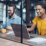 A woman with a headset engages in a VoIP services call while a man works on a laptop at his desk, focused on his phone call.